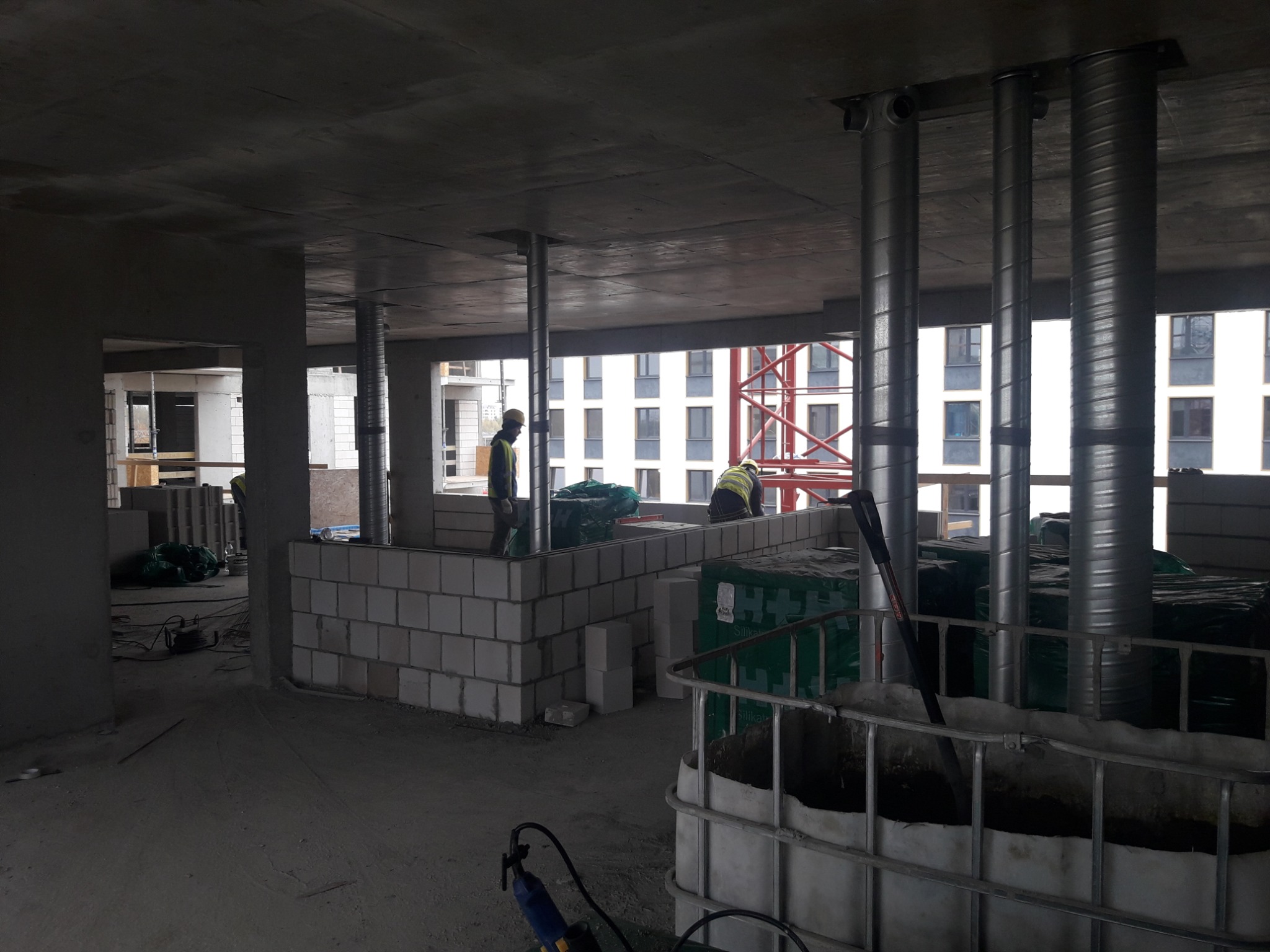 Wnętrze surowego budynku w trakcie budowy, widoczne murowane ściany z betonu komórkowego, stalowe podpory stropu, dwóch robotników w odblaskowych kamizelkach oraz pojemnik IBC z zaprawą.