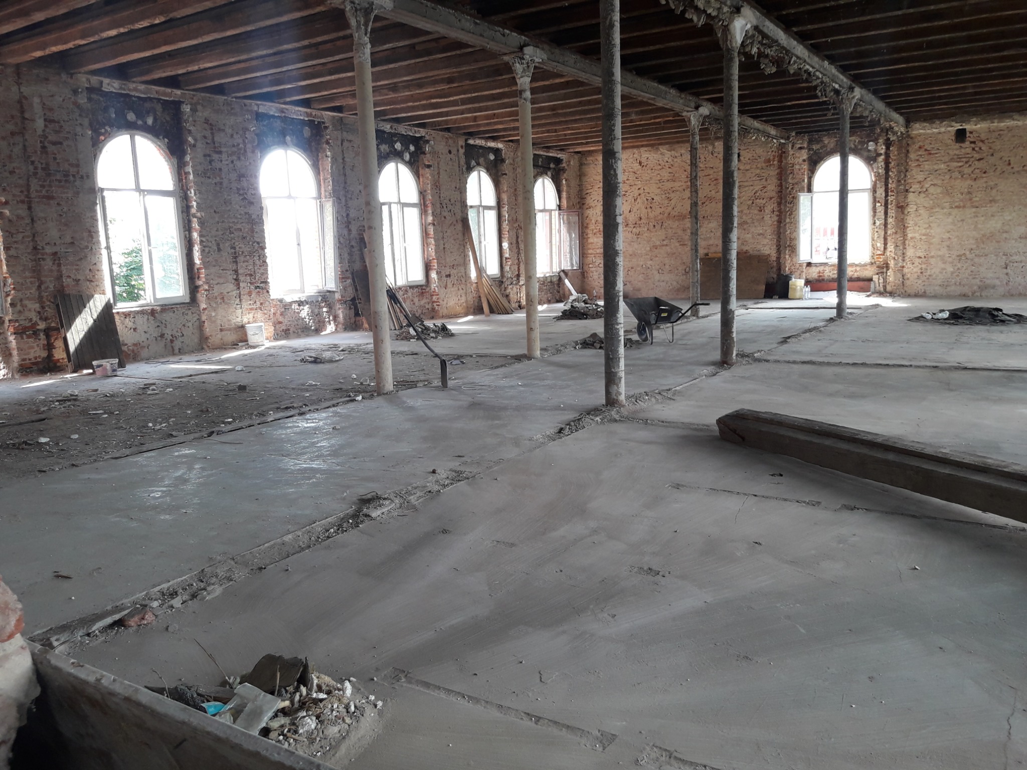 Wnętrze remontowanego budynku z ceglanymi ścianami, kolumnami wspierającymi drewniany strop i świeżo wylaną, szarą posadzką betonową. W tle widoczne okna łukowe i porozrzucane narzędzia.