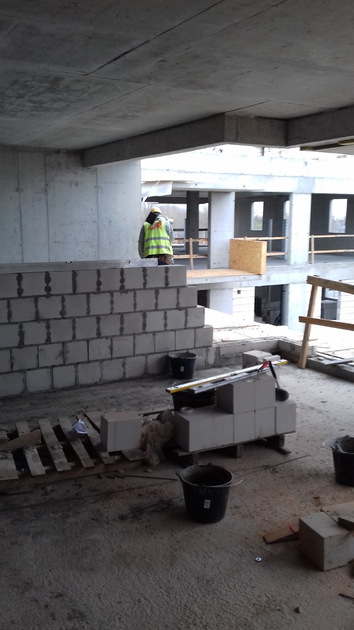 Wnętrze surowego budynku w trakcie budowy, widoczny mur z białych bloczków, pracownik w żółtej kamizelce oraz betonowy strop.