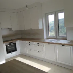 Jasna kuchnia z białymi szafkami, drewnianym blatem i widokiem na zalesione wzgórza za oknem.