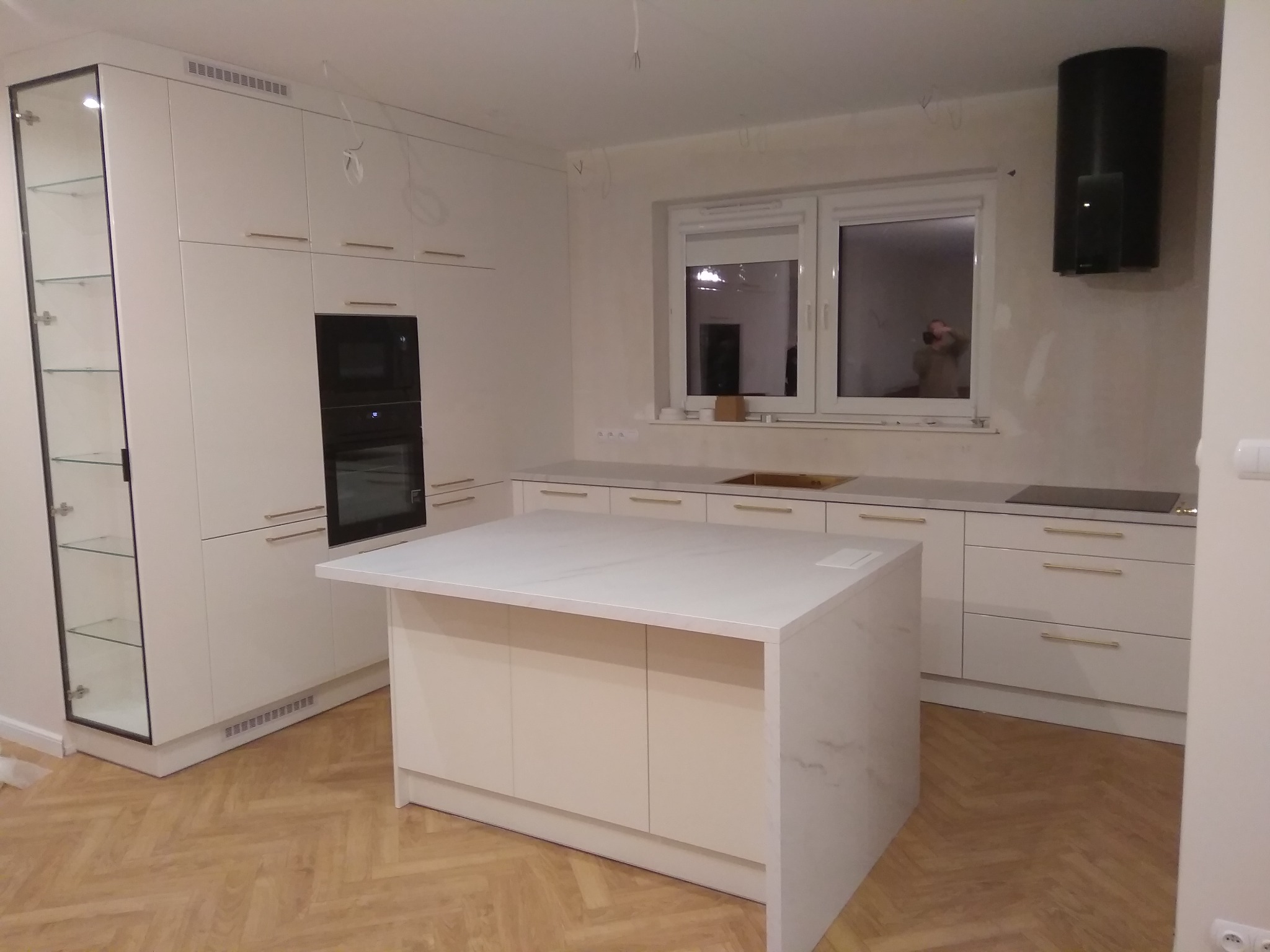 Jasna kuchnia z wyspą, fronty szafek bez uchwytów, złote listwy, czarny piekarnik i okap, podłoga w jodełkę, widoczne przewody elektryczne pod sufitem.