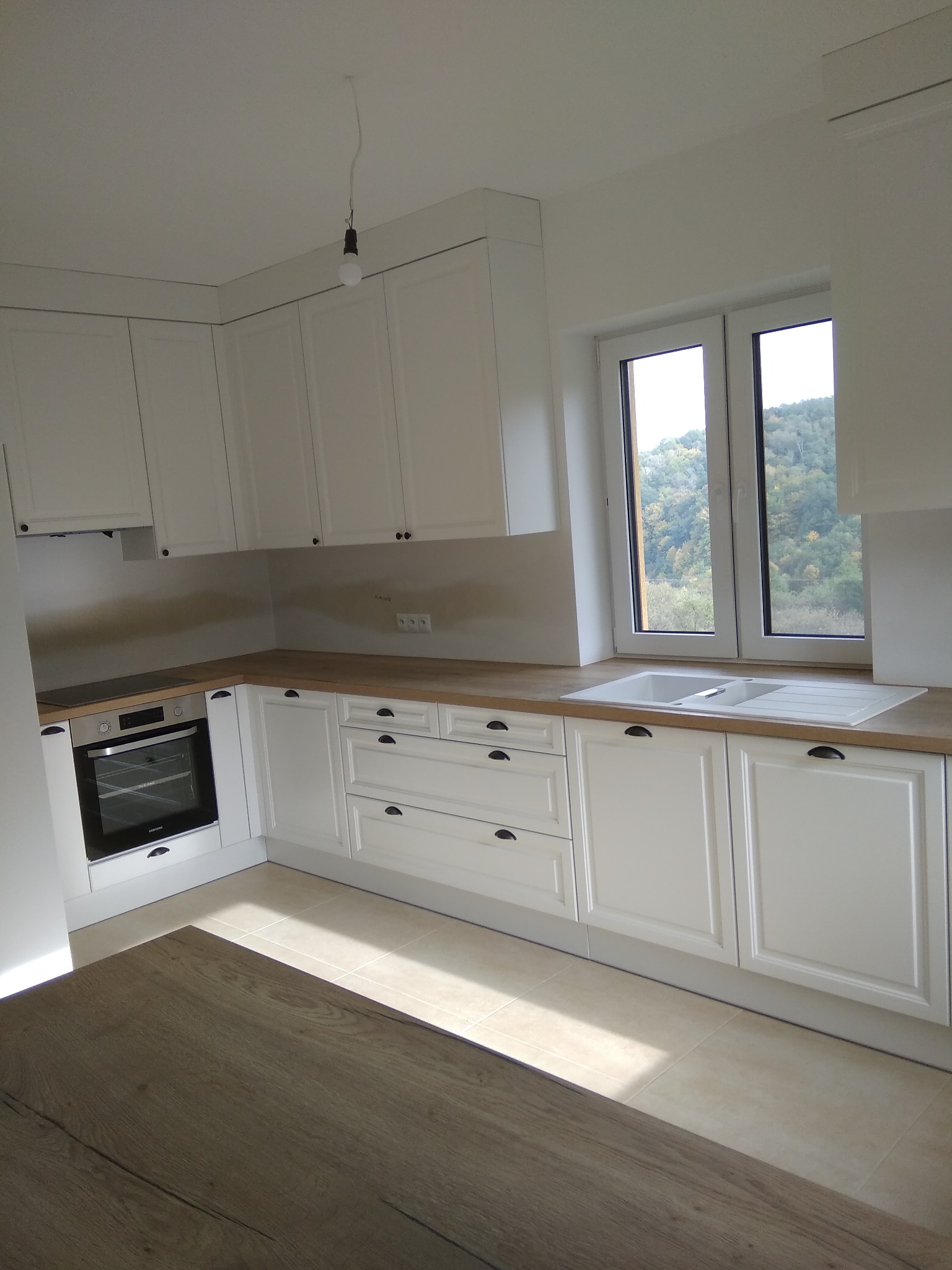 Jasna kuchnia z białymi szafkami, drewnianym blatem i widokiem na zalesione wzgórza za oknem.