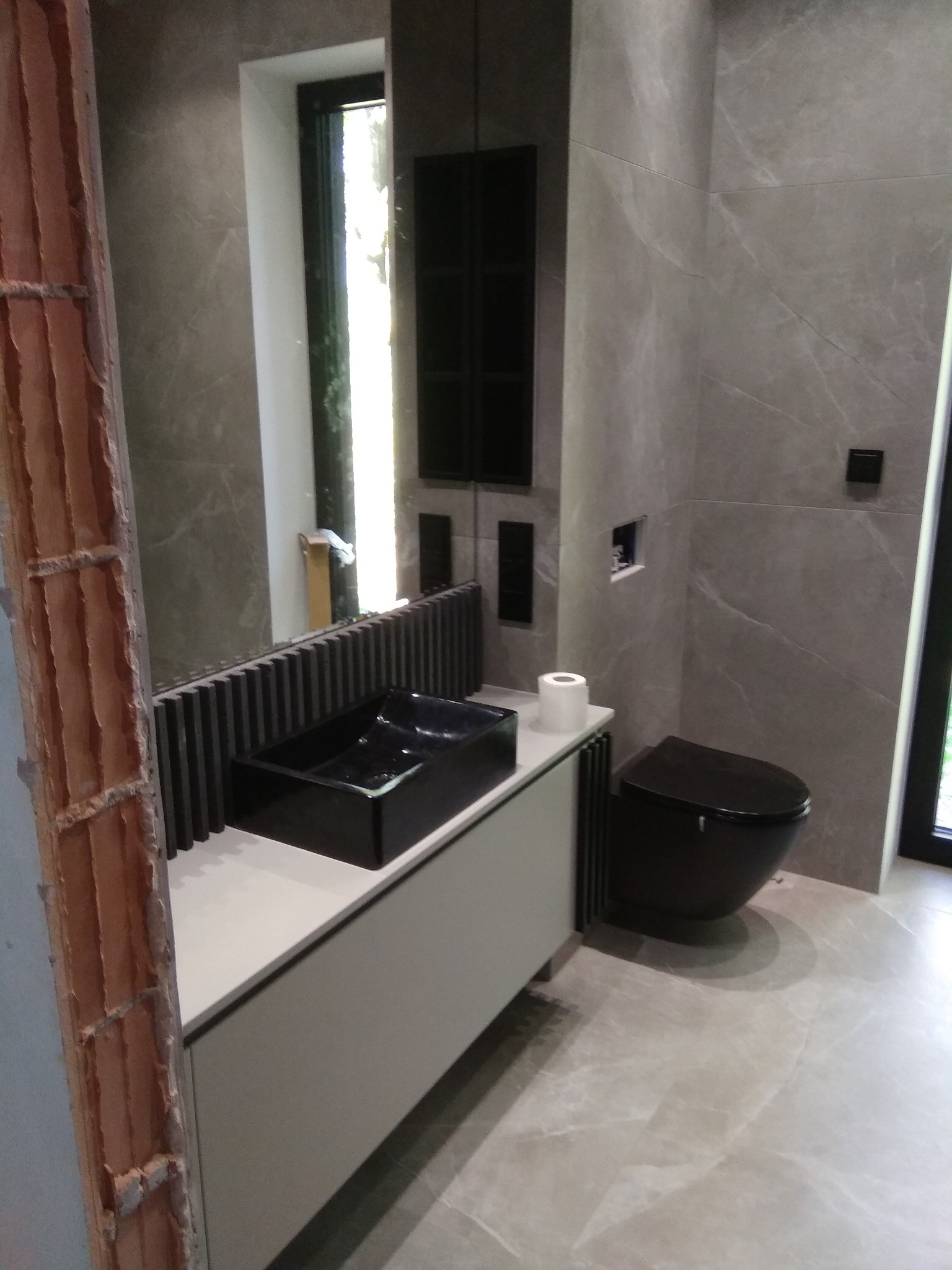 Nowoczesna łazienka w szarościach z czarną umywalką nablatową i toaletą, widoczny fragment surowej ściany z cegły