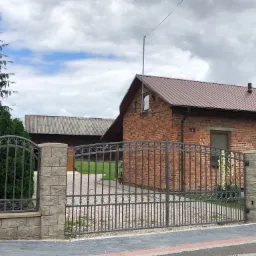 Otwarta, metalowa brama wjazdowa z dekoracyjnymi elementami, osadzona na murowanych słupkach z szarej kostki, prowadząca do ceglanego domu z brązowym dachem.