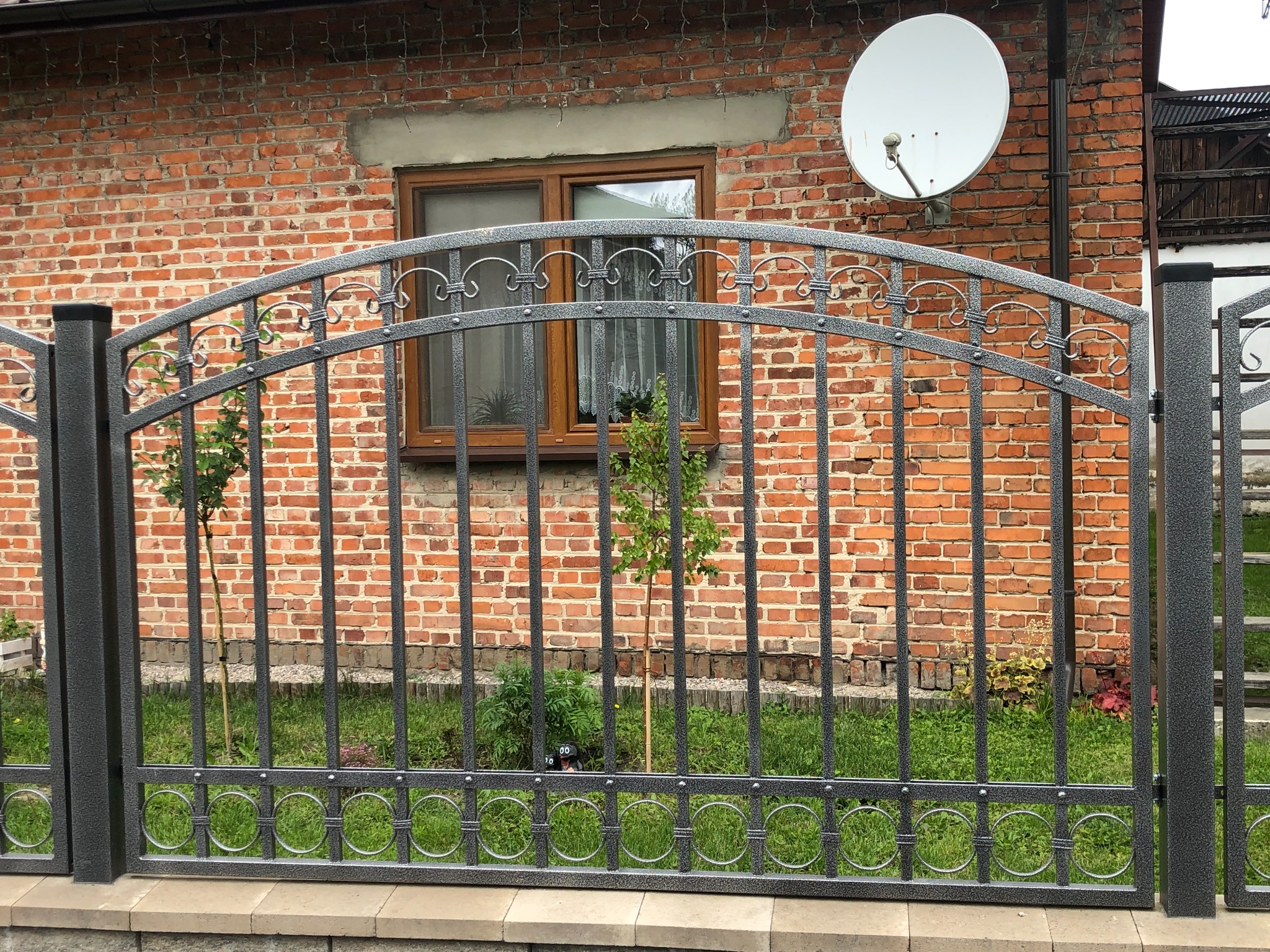 Szare, metalowe ogrodzenie z dekoracyjnymi elementami na tle ceglanego domu z anteną satelitarną.