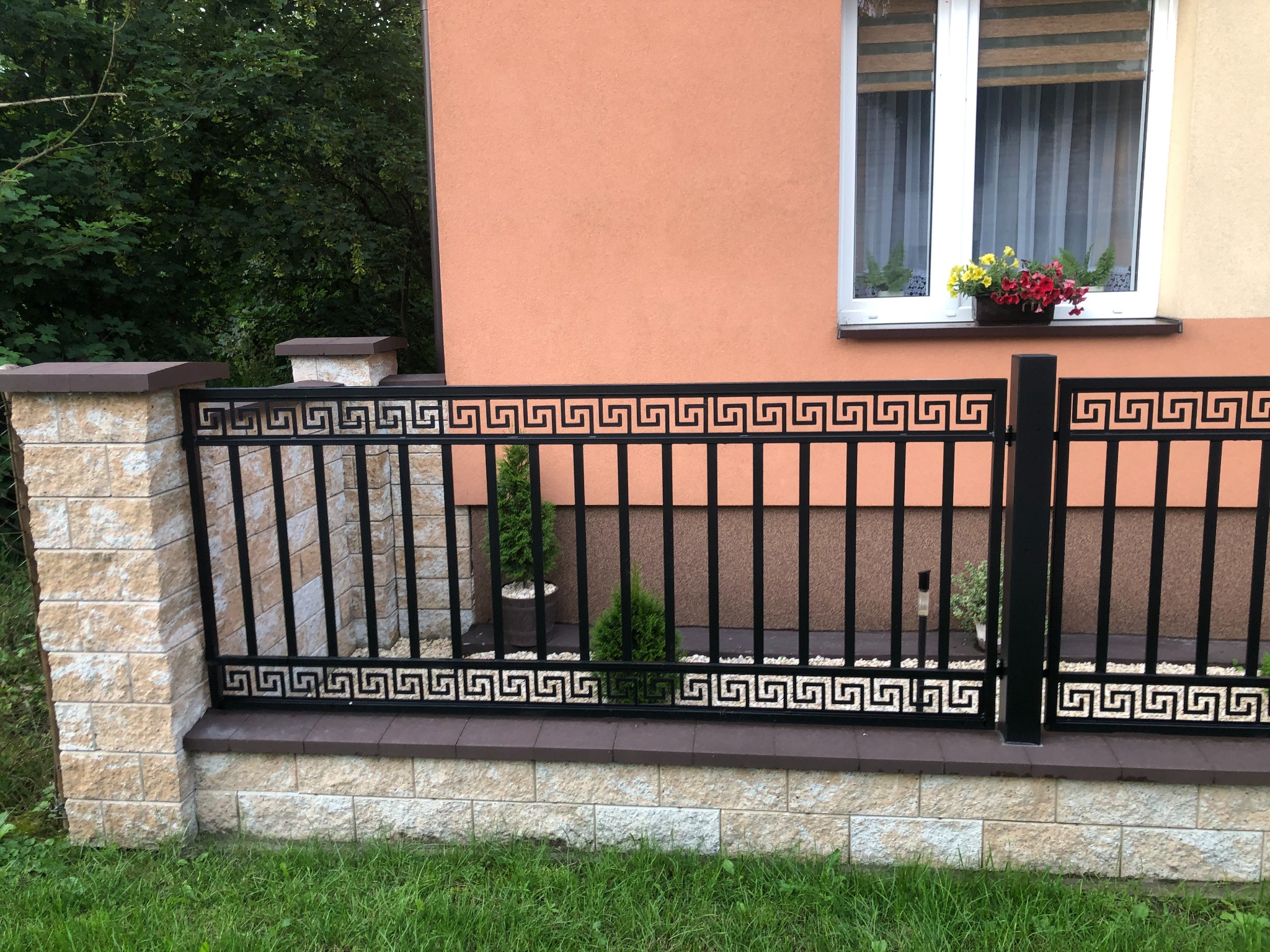 Czarne, metalowe ogrodzenie z motywem greckim, wsparte na słupkach z jasnego kamienia, na tle domu z kwiatami w oknie.