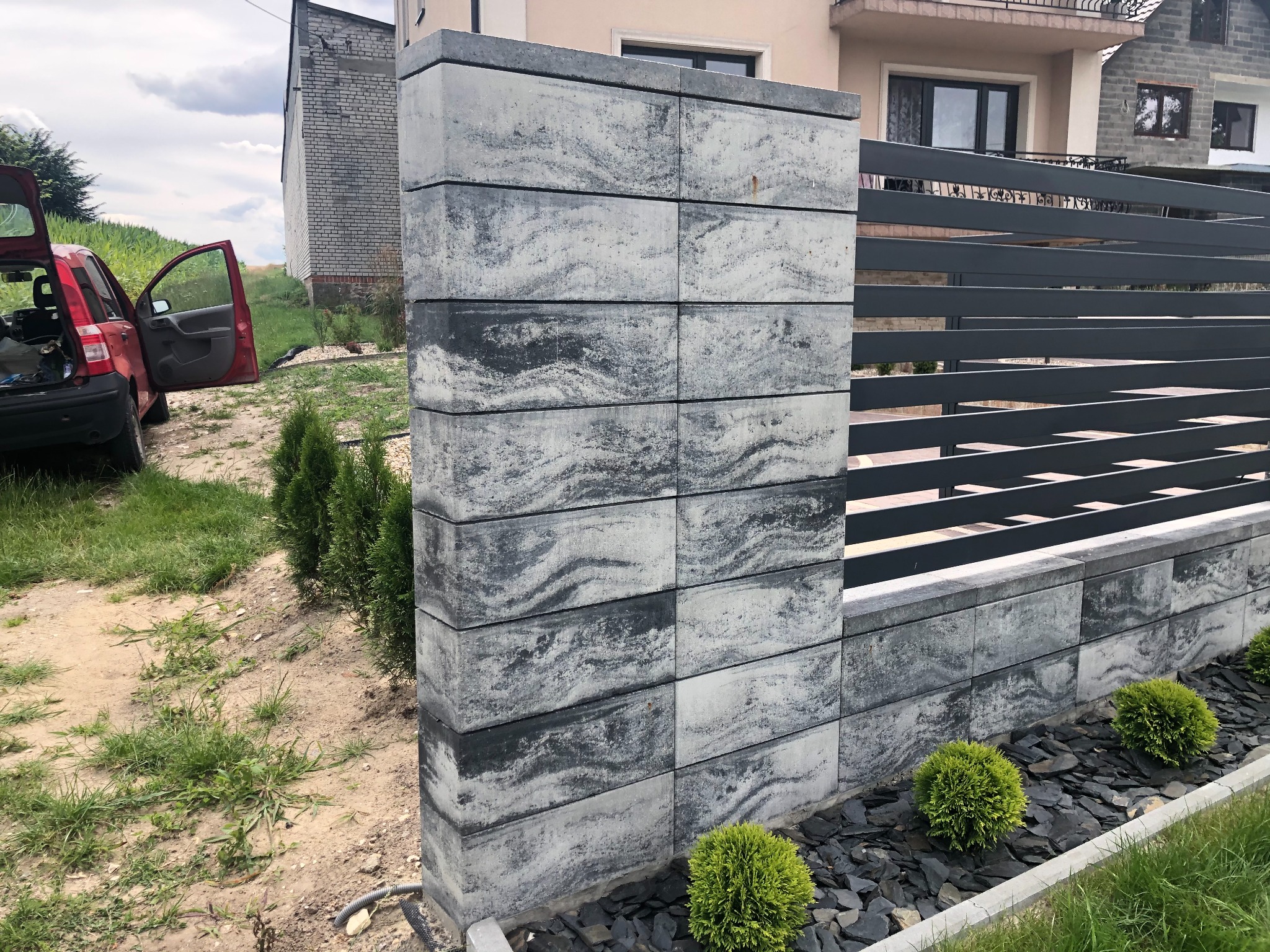 Nowoczesne ogrodzenie z betonowych bloczków w odcieniach szarości, z poziomymi metalowymi przęsłami, otoczone ozdobnym żwirem i niskimi krzewami, z otwartym samochodem w tle.