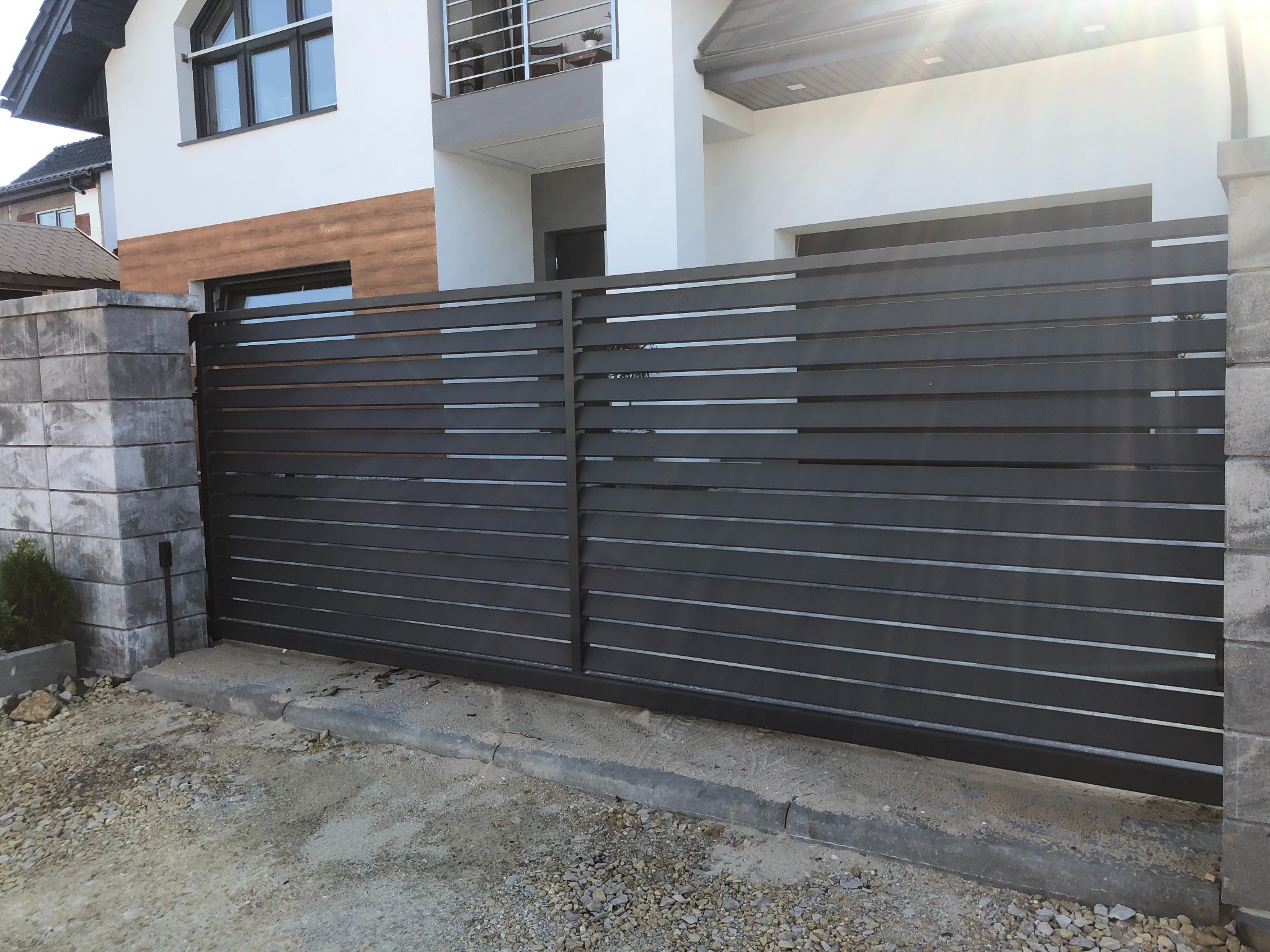 Nowoczesna, przesuwna brama wjazdowa z poziomych, metalowych profili w kolorze antracytowym, zamontowana na posesji z białym domem i szarymi, betonowymi słupkami.