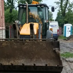 Żółta koparko-ładowarka JCB z brudną łyżką stoi przed budynkiem z ceglaną elewacją, w tle widoczny samochód dostawczy i przenośna toaleta.