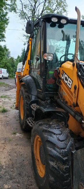 Żółta koparko-ładowarka JCB, lekko zabrudzona, zaparkowana na ziemi; widoczne opony z bieżnikiem, kabina operatora i logo JCB.