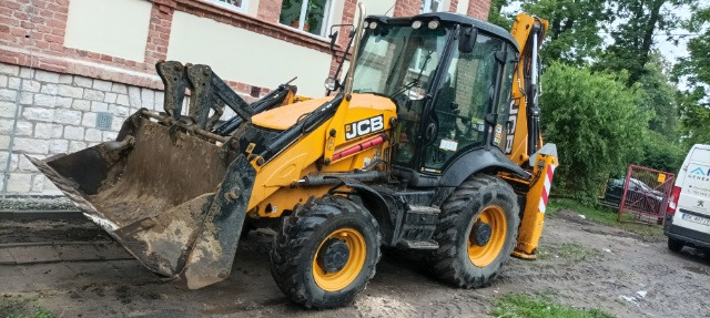 Żółto-czarna koparko-ładowarka JCB z łyżką wypełnioną ziemią, zaparkowana na ubitej ziemi przed budynkiem.