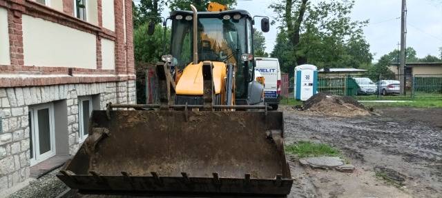 Żółta koparko-ładowarka JCB z brudną łyżką stoi przed budynkiem z ceglaną elewacją, w tle widoczny samochód dostawczy i przenośna toaleta.
