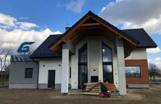 Nowoczesny dom jednorodzinny z dużymi oknami, drewnianą elewacją i elementami wykończeniowymi w trakcie budowy, widoczne palety i sprzęt budowlany przed wejściem.