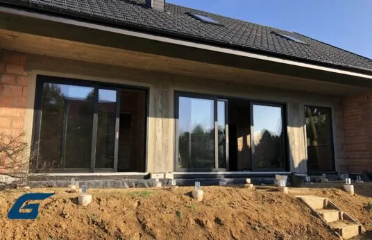 Montaż dużych, przesuwnych okien tarasowych z czarnymi ramami w surowym stanie budynku, widoczne przygotowane podłoże i schody wejściowe.