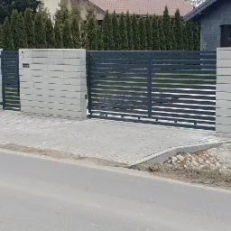 Nowoczesne, poziome przęsła w kolorze grafitowym, osadzone na betonowych słupkach i podmurówce z szarej kostki, tworzące ogrodzenie i bramę wjazdową posesji z widocznym chodnikiem z kostki brukowej...