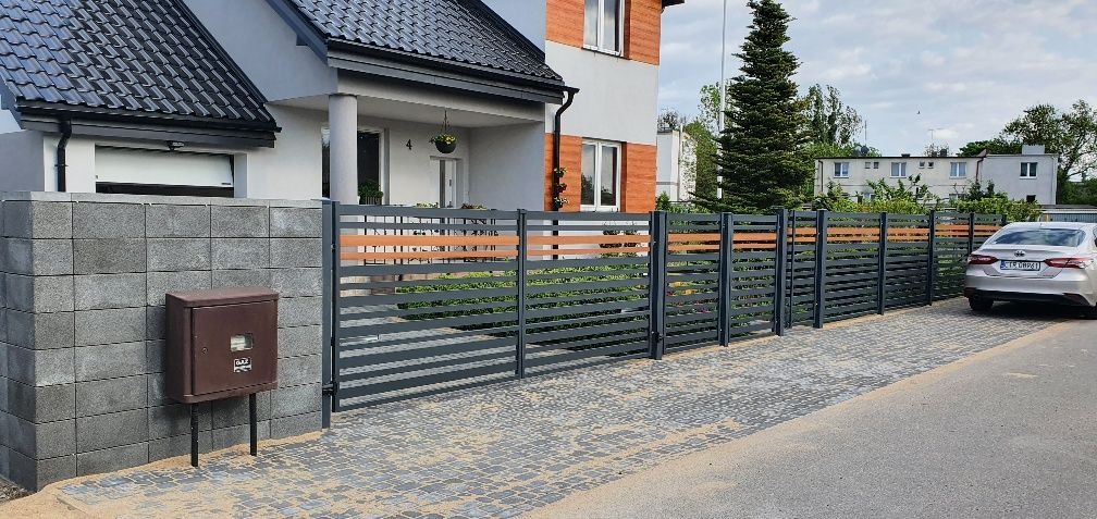 Nowoczesne ogrodzenie panelowe z poziomymi, szarymi przęsłami i drewnianymi akcentami, zamontowane przed domem z szarą kostką brukową i skrzynką pocztową, w tle zaparkowany srebrny samochód.