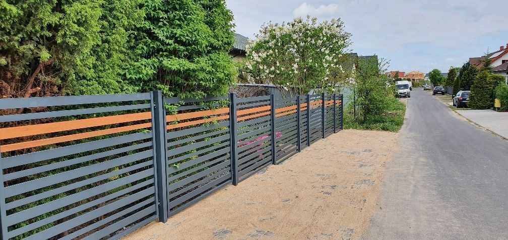 Nowoczesne ogrodzenie panelowe z poziomymi, szarymi przęsłami i wstawkami imitującymi drewno, widoczne z perspektywy ulicy, z zielenią w tle.
