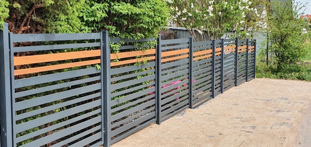 Nowoczesne ogrodzenie panelowe w kolorze grafitowym z poziomymi, drewnopodobnymi wstawkami, widoczne z zewnątrz posesji, z zielenią w tle.