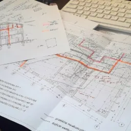 Rozłożone plany architektoniczne z naniesionymi odręcznymi notatkami i kolorowymi oznaczeniami leżące na biurku obok białej klawiatury.
