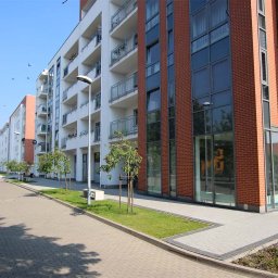 AWiGu_projekt Alina Więcek-Guzińska - Nowoczesny budynek wielorodzinny z balkonami i ceglaną elewacją, widok z poziomu chodnika z kostki brukowej, młode drzewka posadzone wzdłuż budynku.