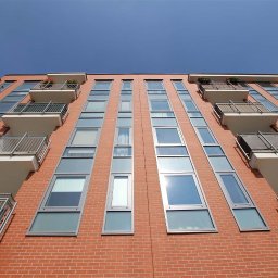 AWiGu_projekt Alina Więcek-Guzińska - Fasada budynku mieszkalnego z cegły z rzędami okien i balkonami z metalowymi balustradami, widok z dołu w słoneczny dzień.