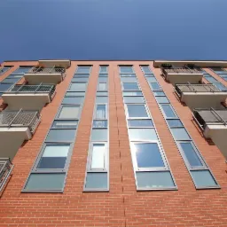 Fasada budynku mieszkalnego z cegły z rzędami okien i balkonami z metalowymi balustradami, widok z dołu w słoneczny dzień.