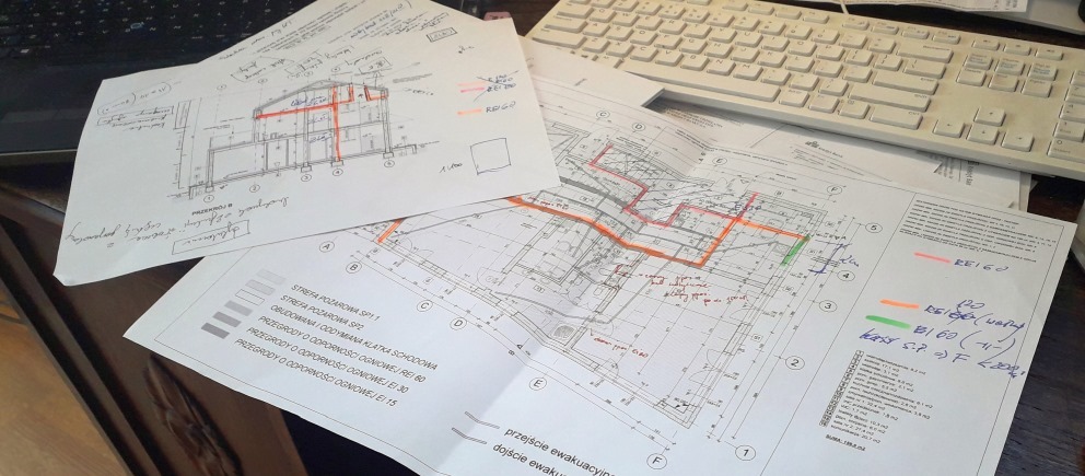 Rozłożone plany architektoniczne z naniesionymi odręcznymi notatkami i kolorowymi oznaczeniami leżące na biurku obok białej klawiatury.