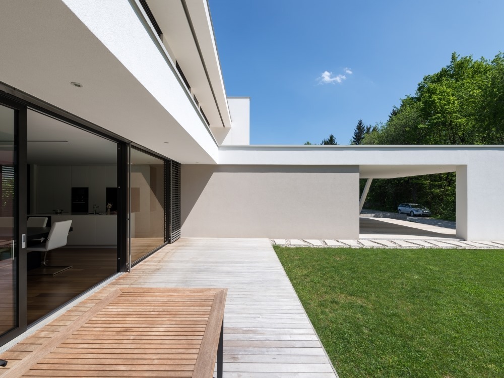 Nowoczesny dom jednorodzinny z drewnianym tarasem, przesuwnymi drzwiami szklanymi i minimalistycznym carportem w tle, otoczony zielonym trawnikiem i drzewami.