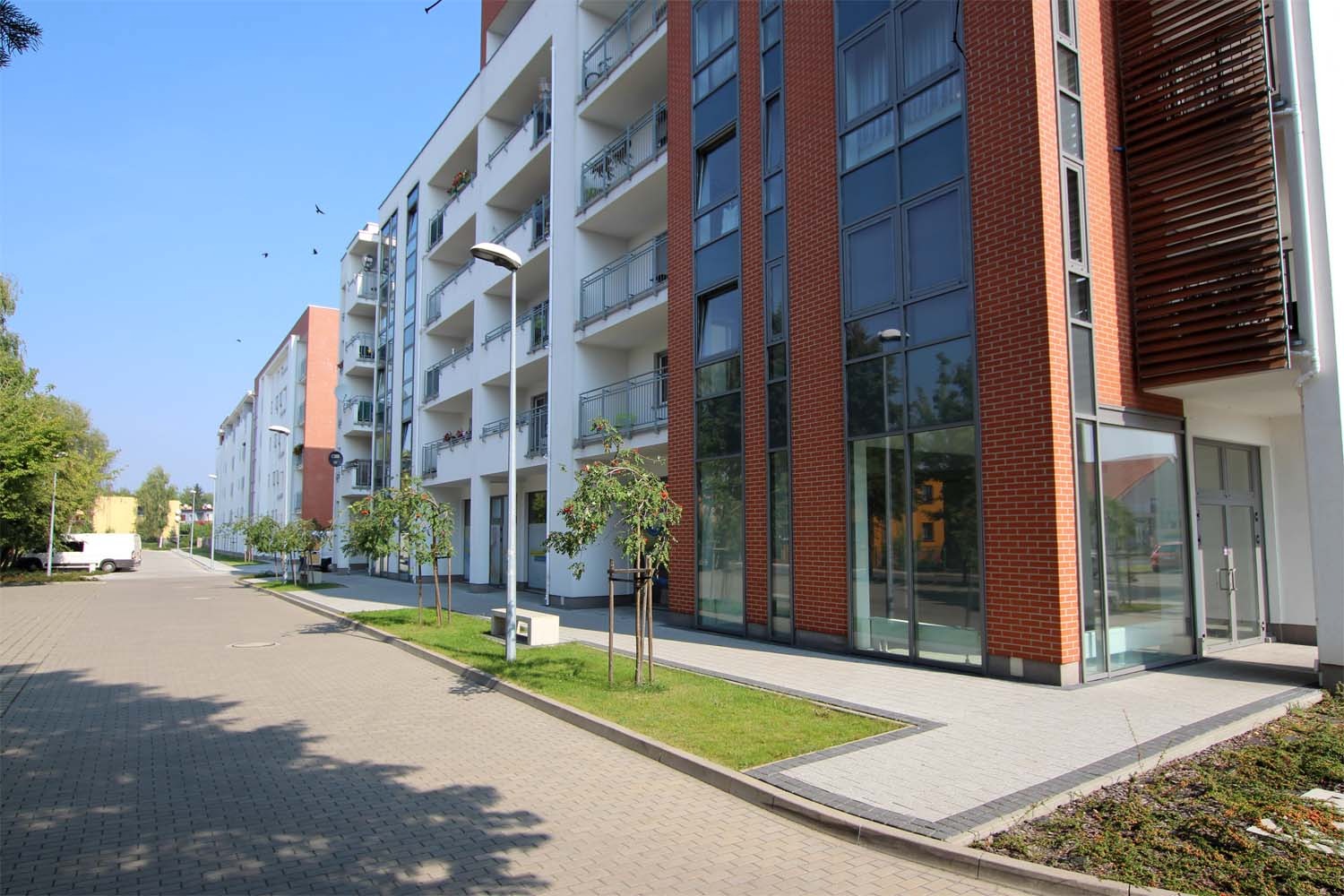 Nowoczesny budynek wielorodzinny z balkonami i ceglaną elewacją, widok z poziomu chodnika z kostki brukowej, młode drzewka posadzone wzdłuż budynku.