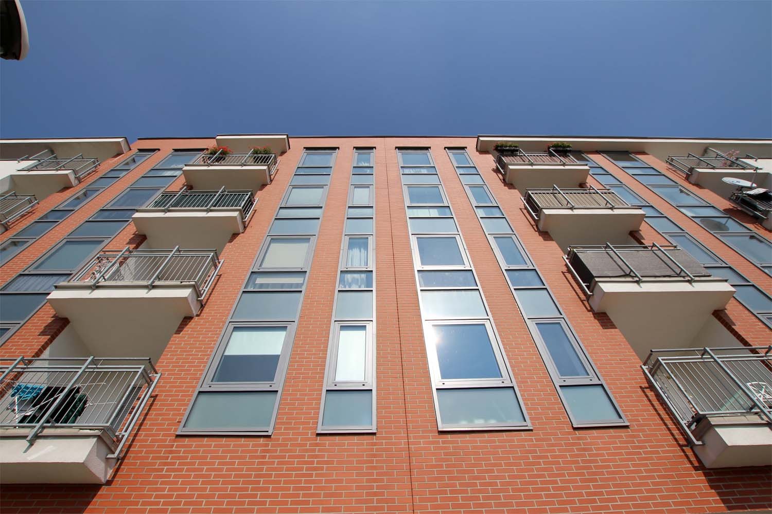 Fasada budynku mieszkalnego z cegły z rzędami okien i balkonami z metalowymi balustradami, widok z dołu w słoneczny dzień.