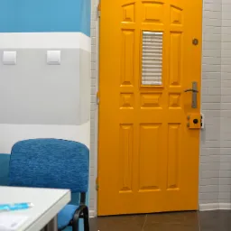 Żółte drzwi z małym okienkiem z żaluzjami w biurze agencji nieruchomości, obok stoją niebieskie krzesła i stolik z ulotką, a pod ścianą leży karton z napisem 'Na sprzedaż' i danymi kontaktowymi biura.