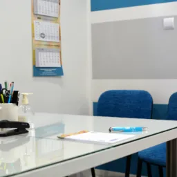 Biuro: Biały stół z szklanym blatem, na nim przybory biurowe, zszywacz, długopis i kalendarz. W tle dwa niebieskie krzesła i kalendarz wiszący na ścianie.