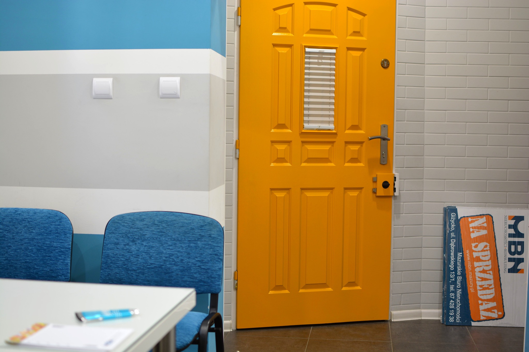 Żółte drzwi z małym okienkiem z żaluzjami w biurze agencji nieruchomości, obok stoją niebieskie krzesła i stolik z ulotką, a pod ścianą leży karton z napisem 'Na sprzedaż' i danymi kontaktowymi biura.