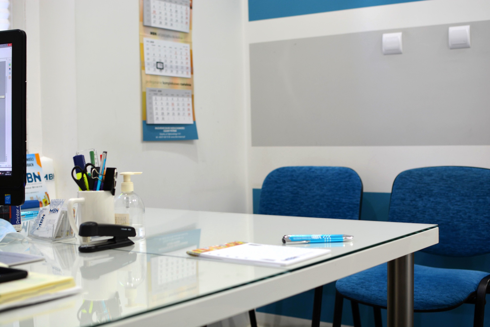 Biuro: Biały stół z szklanym blatem, na nim przybory biurowe, zszywacz, długopis i kalendarz. W tle dwa niebieskie krzesła i kalendarz wiszący na ścianie.