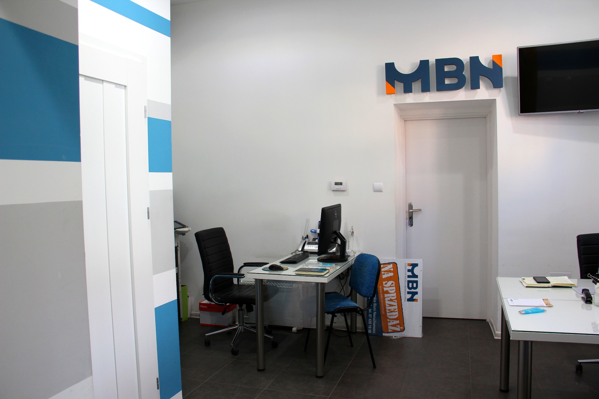 Wnętrze biura nieruchomości z dwoma biurkami, krzesłami, komputerami i logo firmy MBN na ścianie. Widoczny plakat reklamowy z napisem 'NA SPRZEDAŻ'.