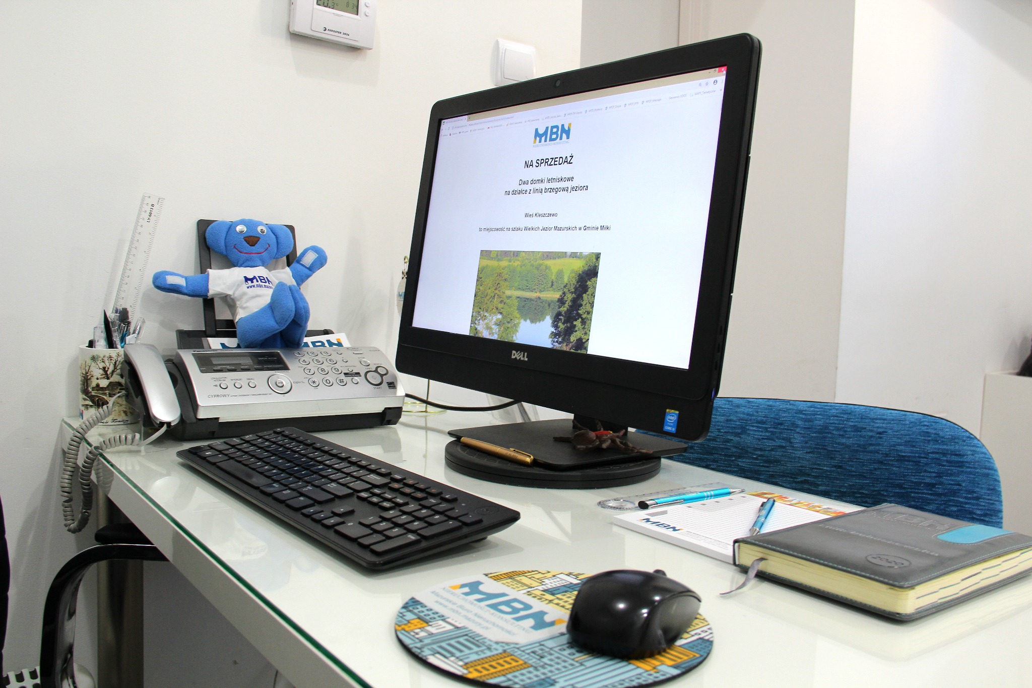 Biuro z komputerem wyświetlającym ofertę sprzedaży domków letniskowych, telefonem, klawiaturą, myszką na podkładce z logo firmy MBN i pluszowym misiem w koszulce MBN siedzącym na telefonie.