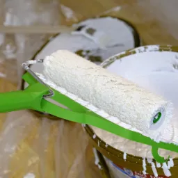 Wałek malarski z zieloną rączką zanurzony w wiadrze z białą farbą, widoczna folia ochronna w tle.