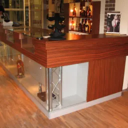 Bar recepcyjny z blatem z drewna egzotycznego, szklanymi elementami ekspozycyjnymi i metalowymi wspornikami, w tle regał z alkoholem i pracownik obsługi.