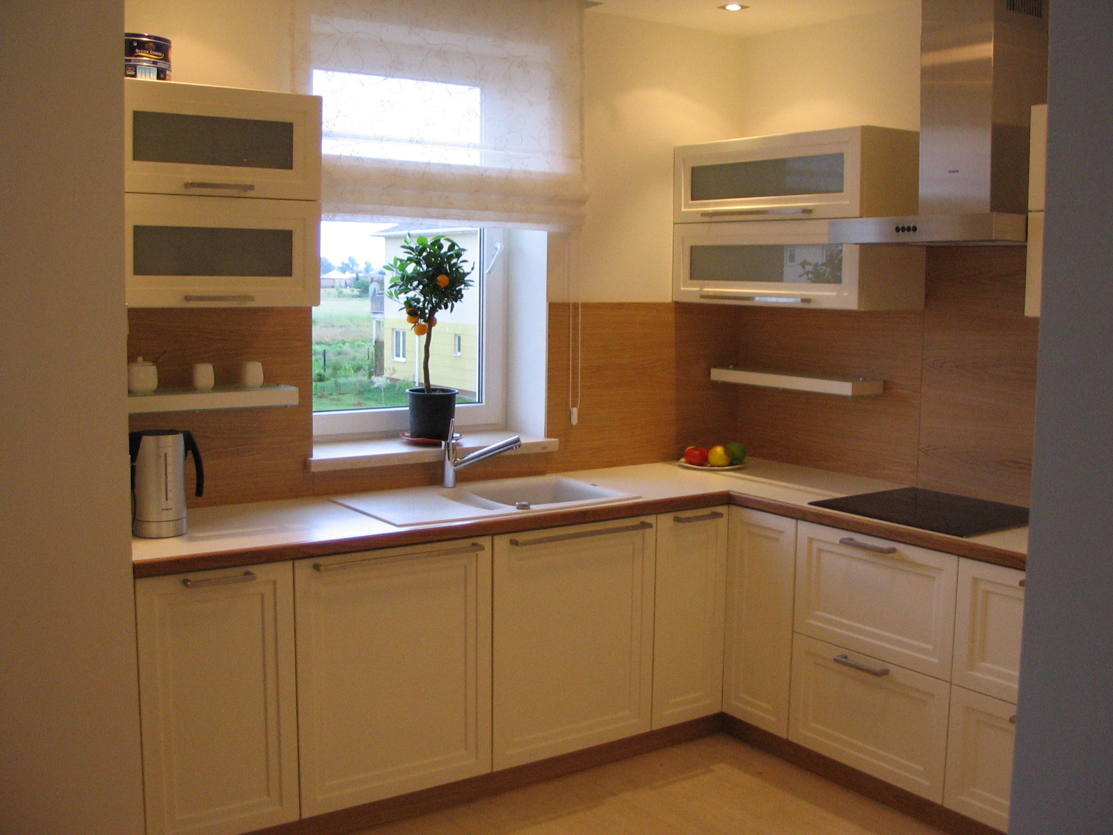 Jasna kuchnia w stylu klasycznym z drewnianymi akcentami, wyposażona w szafki z przeszkleniami, nowoczesny okap i małe drzewko cytrusowe w oknie.