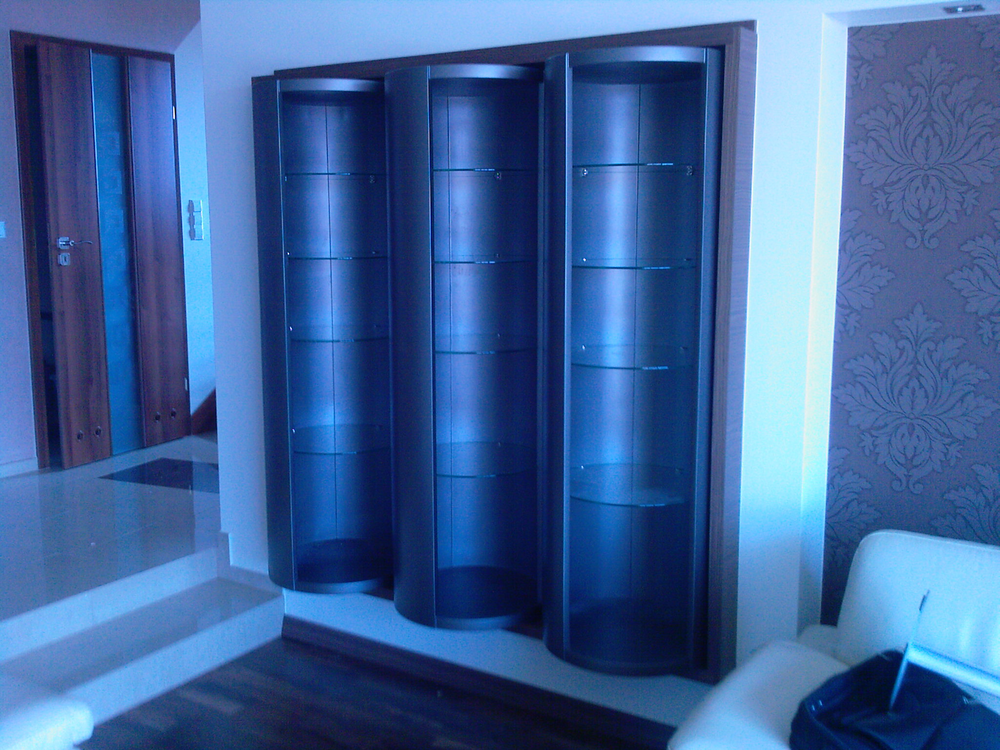 Trzy wysokie, ciemne witryny z półkami, umieszczone obok schodów i ściany z wzorzystą tapetą w pomieszczeniu z chłodnym oświetleniem.