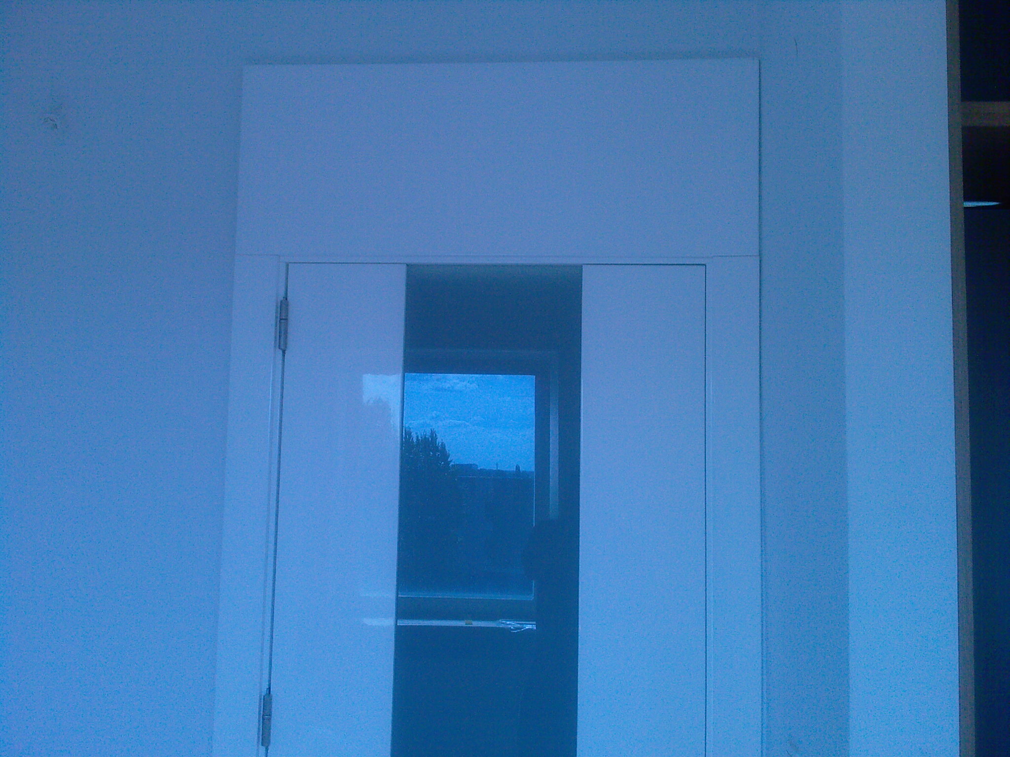 Białe drzwi wewnętrzne z prostokątną szybą, zamontowane w białej ścianie, widok na zewnątrz przez szybę.