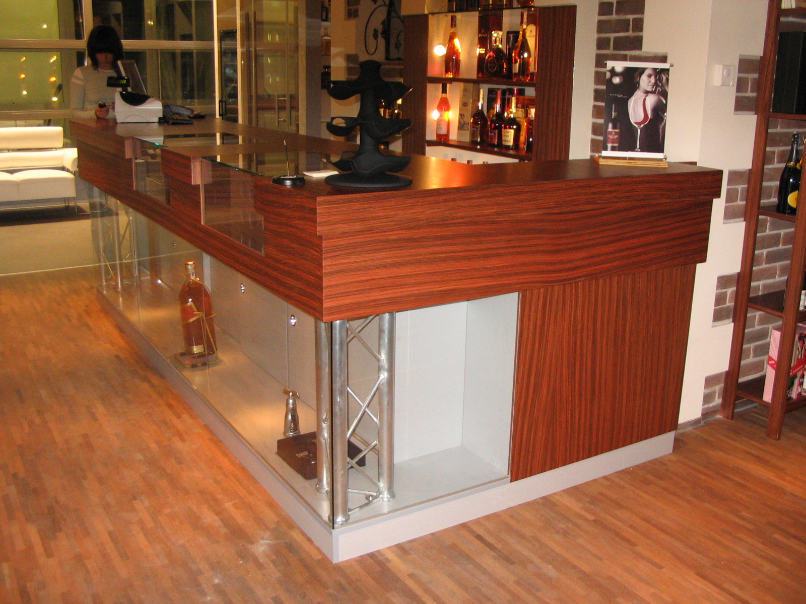 Bar recepcyjny z blatem z drewna egzotycznego, szklanymi elementami ekspozycyjnymi i metalowymi wspornikami, w tle regał z alkoholem i pracownik obsługi.