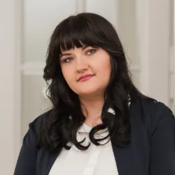 Portret uśmiechniętej kobiety z ciemnymi włosami w białej bluzce i granatowej marynarce, opierającej się o białą ścianę z przeszklonymi drzwiami w tle.