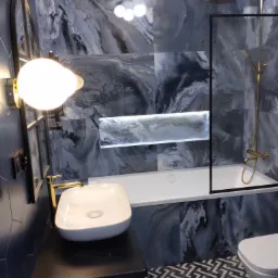 Nowoczesna łazienka z marmurowymi płytkami w odcieniach szarości i bieli, z podświetlaną niszą nad wanną, złotą armaturą i geometryczną podłogą.