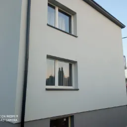 Świeżo otynkowana elewacja domu jednorodzinnego w odcieniach szarości, z widocznymi dwoma oknami i rynną, uchwycona pod kątem z poziomu gruntu.