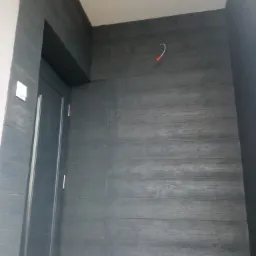 Elewacja budynku wykończona panelami imitującymi drewno w odcieniach szarości, z widoczną ramą drzwi i wystającym kablem ze ściany.