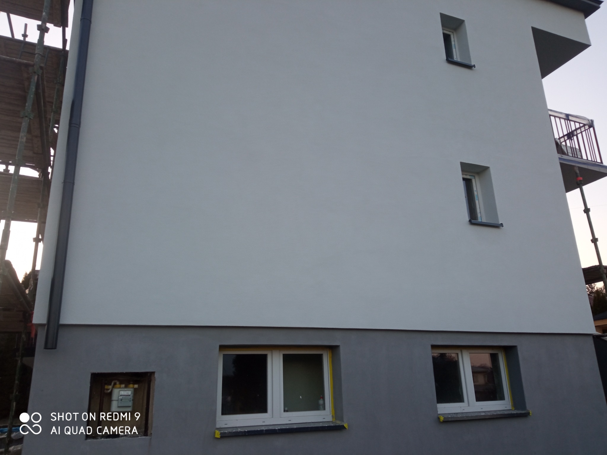 Świeżo otynkowana elewacja budynku z widocznymi rusztowaniami, rurą spustową i oknami zabezpieczonymi taśmą malarską, z szarą podmurówką i jasną resztą elewacji.