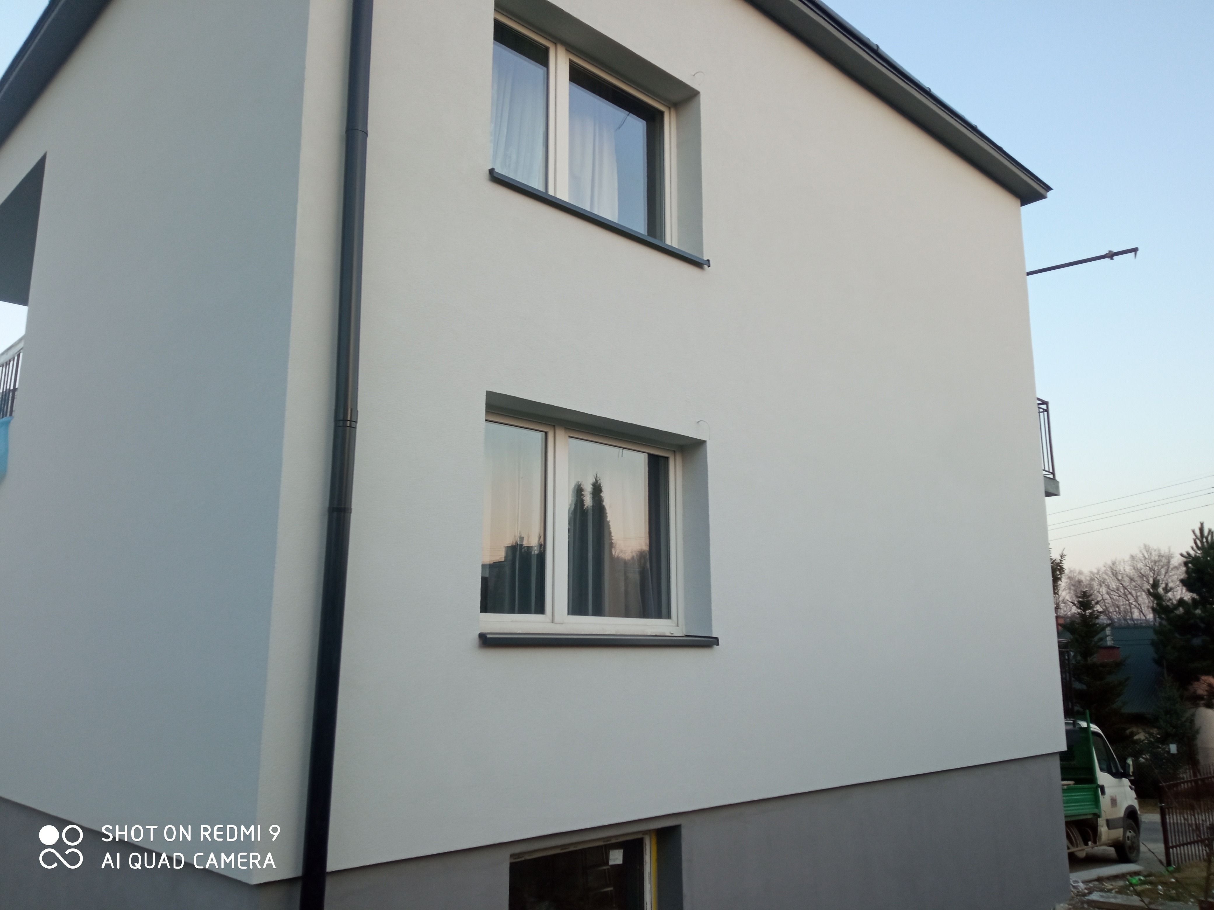 Świeżo otynkowana, dwukondygnacyjna elewacja domu w jasnym odcieniu szarości, z widocznymi oknami z ciemnymi ramami, rynną spustową i fragmentem balkonu w tle