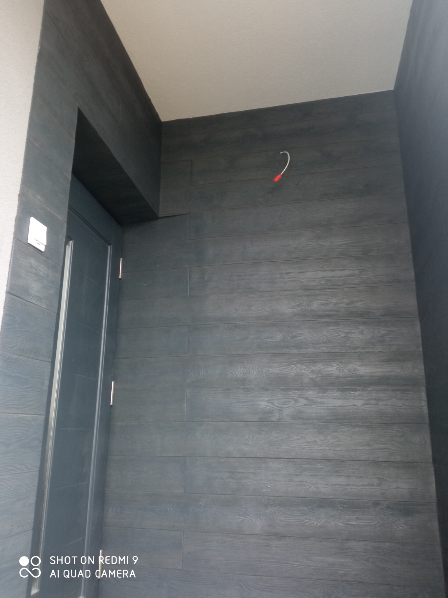 Elewacja budynku wykończona panelami imitującymi drewno w odcieniach szarości, z widoczną ramą drzwi i wystającym kablem ze ściany.