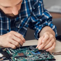 Mężczyzna w niebieskiej koszuli w kratę instaluje moduł pamięci RAM w płycie głównej laptopa, widoczne elementy chłodzenia i inne podzespoły.